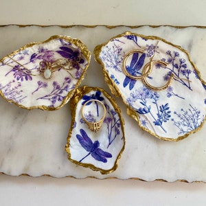 May include: Three white ceramic oyster shell dishes with blue and gold dragonfly designs. The dishes are perfect for holding jewelry, rings, earrings, or other small items.