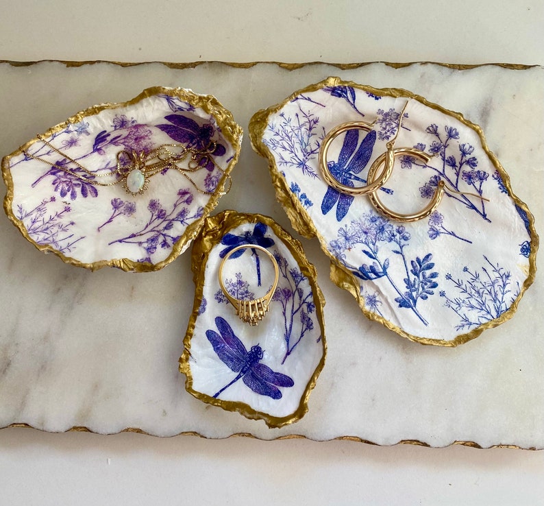 May include: Three white ceramic oyster shell dishes with blue and gold dragonfly designs. The dishes are perfect for holding jewelry, rings, earrings, or other small items.
