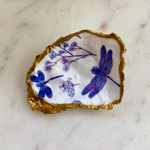 May include: A white oyster shell with a gold rim, decorated with a blue and purple dragonfly and floral designs.