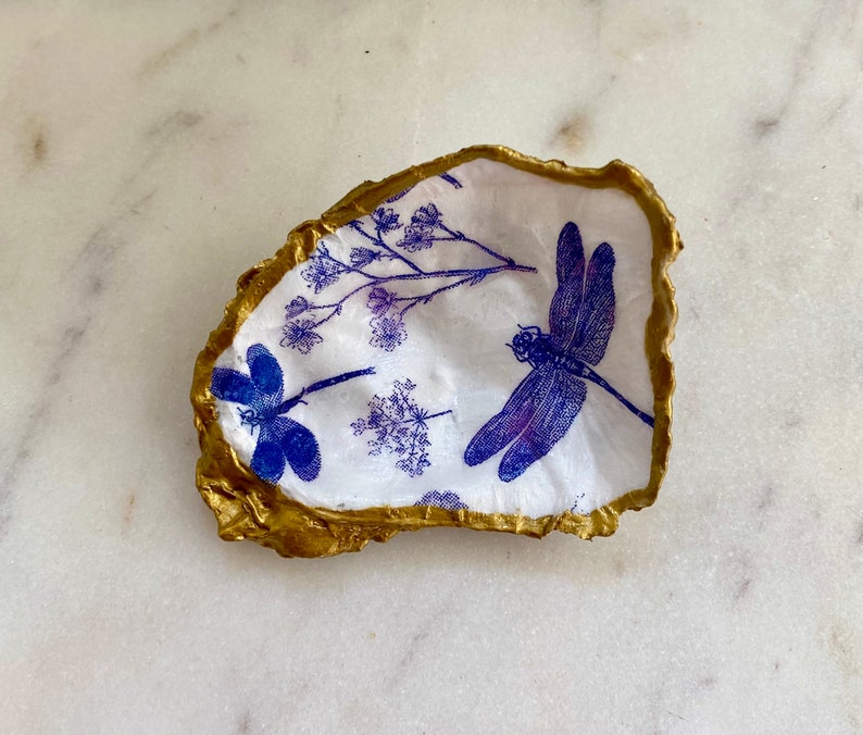 May include: A white oyster shell with a gold rim, decorated with a blue and purple dragonfly and floral designs.