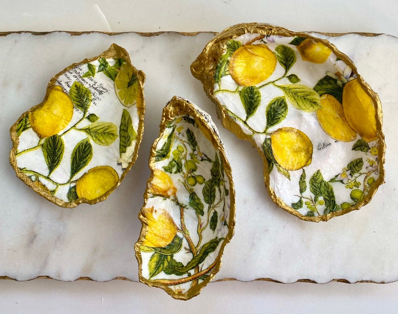 May include: Three golden oyster shells with a lemon and leaf design. The shells are on a white marble surface.