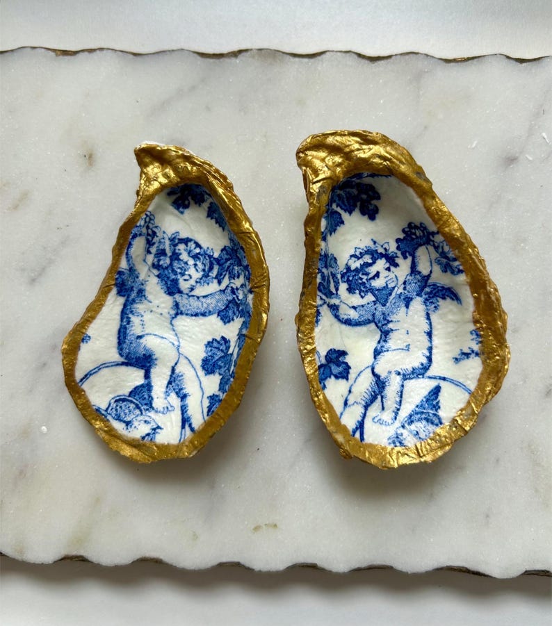 Pu&ograve; includere: Un paio di orecchini a forma di conchiglia di ostrica con un design di cherubino blu e bianco e bordi dorati.