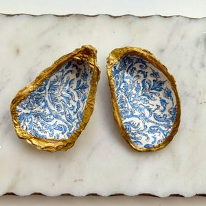 May include: Two gold-rimmed oyster shells with a blue and white floral pattern. The shells are on a white marble surface.