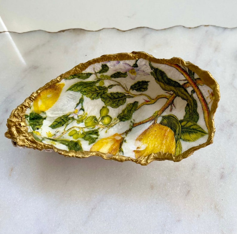 May include: A white ceramic oyster shell dish with a gold rim. The inside of the dish is decorated with a lemon tree design featuring yellow lemons, green leaves, and white flowers.