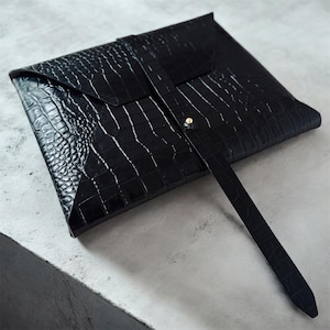 May include: Black faux-leather envelope-style clutch with a crocodile skin texture. The clutch has a flap closure secured by a strap and a gold-colored button. The strap extends from the top and wraps around the front.