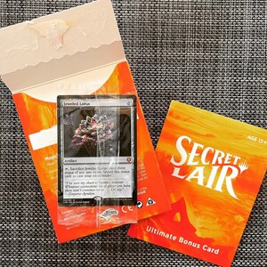 May include: A Secret Lair Ultimate Bonus Card featuring a Magic: The Gathering card, Jeweled Lotus, in a sealed plastic bag. The card is black with a colourful image of a lotus flower. The card is surrounded by a bright orange background with the text "Secret Lair" and "Ultimate Bonus Card".