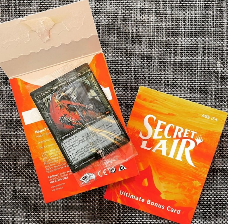 May include: A Secret Lair Ultimate Bonus Card featuring a black and white illustration of a creature with a red background. The card is in a clear plastic sleeve and is surrounded by a bright orange background with the text "Secret Lair" and "Ultimate Bonus Card".