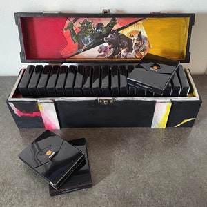 Puede incluir: Una caja de madera con una ilustración colorida de una escena de fantasía en la tapa. La caja está abierta y llena de barajas de cartas negras. Las barajas de cartas tienen un logotipo dorado en la parte delantera.