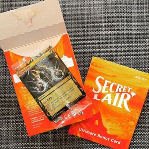 May include: A Secret Lair Ultimate Bonus Card featuring a black and gold Magic: The Gathering card with the text "Dark Ritual!" and "Instant". The card is in a plastic sleeve and is on top of a red and orange card with the text "Secret Lair" and "Ultimate Bonus Card".