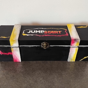May include: A black wooden box with yellow, white, and pink painted stripes. The box has a brass latch and the words "JUMP START" are painted on the lid in white and red.