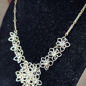 May include: A gold-tone metal necklace featuring a series of interconnected openwork flower motifs. Each floral element is accented with a clear, round-cut rhinestone at its center. The chain consists of textured links leading to the floral centerpiece.
