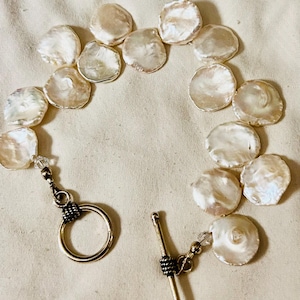 May include: A bracelet featuring large, flat, iridescent pearls. The pearls are strung together, forming a circular shape. The bracelet has a silver-colored clasp with a toggle closure. The pearls have a creamy white color with hints of pink and gold.