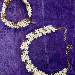 May include: A gold-tone metal jewelry set consisting of a bracelet and a necklace. Both pieces feature a repeating pattern of white enamel flowers with small clear rhinestones at their centers, connected by gold-tone metal links and chains.