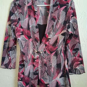 Könnte beinhalten: Ein gemusterter Blazer in Rosa-, Schwarz- und Weißtönen mit einem einzigartigen blattartigen Design. Der Blazer hat Dreiviertelärmel und einen dekorativen Verschluss an der Taille. Das Kleidungsstück hängt an einem weißen Kleiderbügel.