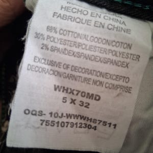 K&ouml;nnte beinhalten: Nahaufnahme eines wei&szlig;en Etiketts mit Text in Englisch, Spanisch und Franz&ouml;sisch. Das Etikett lautet "HECHO EN CHINA" und "FABRIQUE EN CHINE". Es gibt die Stoffzusammensetzung an: 68 % Baumwolle, 30 % Polyester und 2 % Elasthan. Das Etikett hat auch den Produktcode WHX70MD 5 X 32.