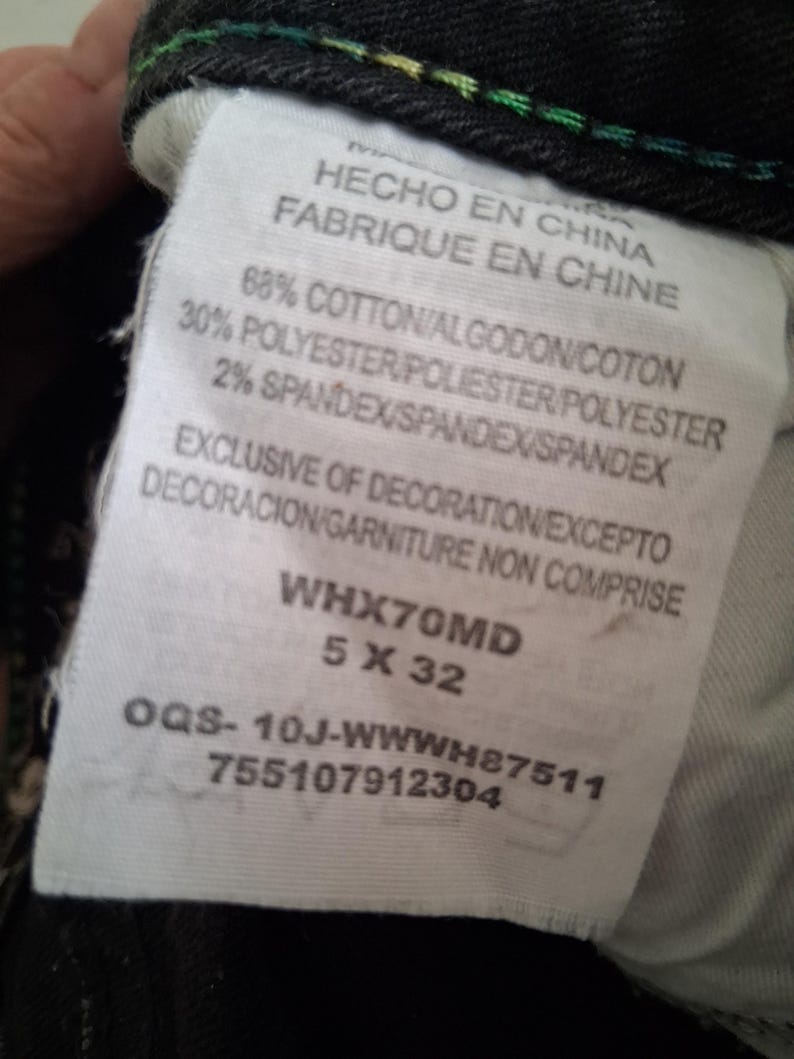 K&ouml;nnte beinhalten: Nahaufnahme eines wei&szlig;en Etiketts mit Text in Englisch, Spanisch und Franz&ouml;sisch. Das Etikett lautet "HECHO EN CHINA" und "FABRIQUE EN CHINE". Es gibt die Stoffzusammensetzung an: 68 % Baumwolle, 30 % Polyester und 2 % Elasthan. Das Etikett hat auch den Produktcode WHX70MD 5 X 32.