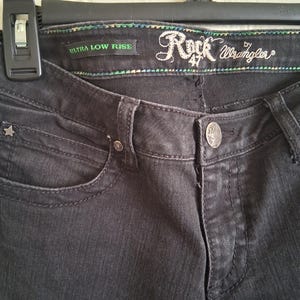 K&ouml;nnte beinhalten: Nahaufnahme einer schwarzen Jeans mit den Worten "Rock 47 Wrangler" auf dem Bund gestickt. Die Jeans hat eine sternf&ouml;rmige Niete und die Worte "Ultra Low Rise" sind sichtbar.