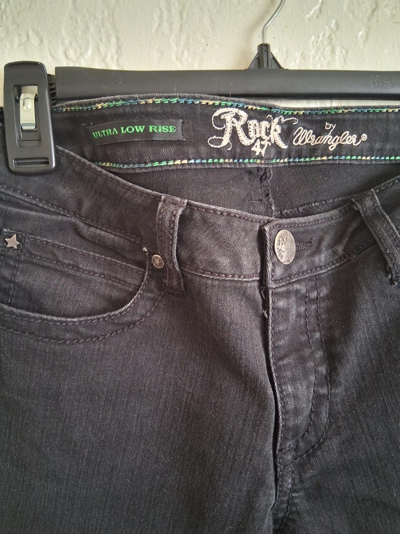 K&ouml;nnte beinhalten: Nahaufnahme einer schwarzen Jeans mit den Worten "Rock 47 Wrangler" auf dem Bund gestickt. Die Jeans hat eine sternf&ouml;rmige Niete und die Worte "Ultra Low Rise" sind sichtbar.