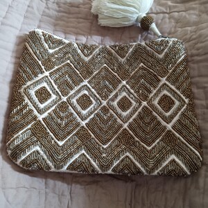 May include: A white beaded clutch with a gold geometric pattern and a white tassel.
