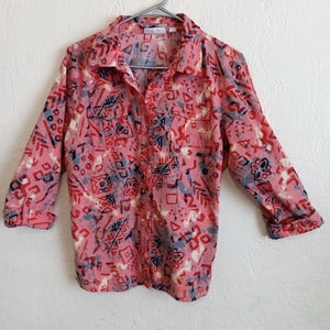 May include: A pink button-up shirt with a geometric pattern in shades of red, blue, and white. The shirt has a collar and long sleeves.