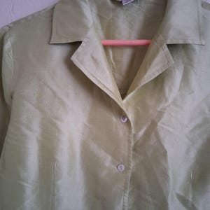 May include: A light green button-down shirt with a collar, hanging on a pink hanger. The shirt has a silky texture and is buttoned up. The fabric appears to have a subtle sheen.