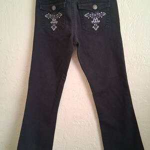 K&ouml;nnte beinhalten: Schwarze Jeans mit Strassverzierungen auf den Ges&auml;&szlig;taschen. Die Jeans haben ein klassisches F&uuml;nf-Taschen-Design, einen Knopf- und Rei&szlig;verschluss und einen leichten Schlag am Beinabschluss.