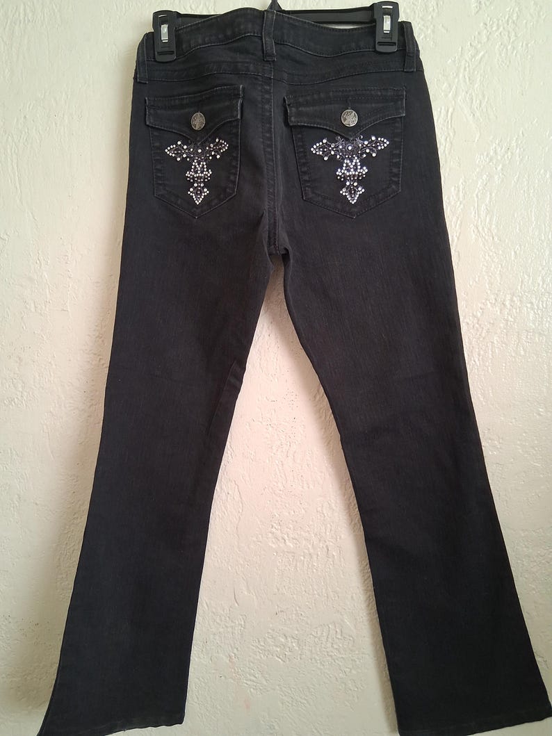 K&ouml;nnte beinhalten: Schwarze Jeans mit Strassverzierungen auf den Ges&auml;&szlig;taschen. Die Jeans haben ein klassisches F&uuml;nf-Taschen-Design, einen Knopf- und Rei&szlig;verschluss und einen leichten Schlag am Beinabschluss.