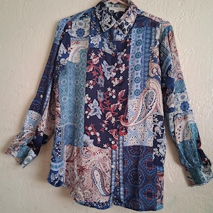 May include: A long-sleeved shirt with a patchwork design in shades of blue, burgundy, and white. The shirt features paisley, floral, and geometric patterns. It is buttoned up and hanging on a pink hanger. The shirt's design is a mix of different patterns.
