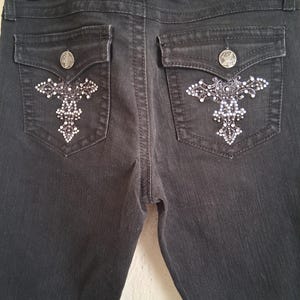K&ouml;nnte beinhalten: Dunkelgraue Jeans mit dekorativen kreuzf&ouml;rmigen Verzierungen auf den Ges&auml;&szlig;taschen. Jede Tasche hat einen silbernen Knopf und ein funkelndes, strassartiges Design. Die Jeans haben ein klassisches F&uuml;nf-Taschen-Design.