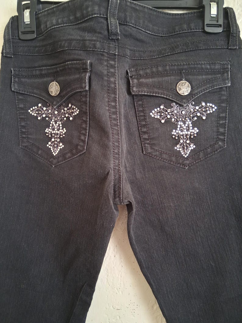 K&ouml;nnte beinhalten: Dunkelgraue Jeans mit dekorativen kreuzf&ouml;rmigen Verzierungen auf den Ges&auml;&szlig;taschen. Jede Tasche hat einen silbernen Knopf und ein funkelndes, strassartiges Design. Die Jeans haben ein klassisches F&uuml;nf-Taschen-Design.
