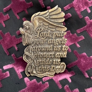 May include: A bronze-colored angel pin with praying hands and large wings. The pin has the words "Lord, put your angels around us to protect and guide us this Day" in a decorative font. The pin is on a black background with a repeating pattern of maroon shapes.
