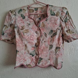 Puede incluir: Una blusa vintage de manga corta con un estampado floral de rosas rosas, verdes y beiges. La blusa tiene un borde festoneado y se abotona en la parte delantera. El ribete es de un rosa más oscuro.