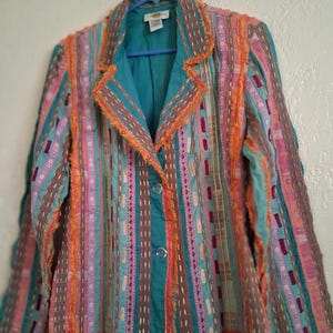 Puede incluir: Una colorida blazer con rayas verticales en tonos turquesa, rosa, naranja y marrón. La chaqueta tiene un forro turquesa y se abrocha con dos botones. El cuello y los bordes están adornados con flecos naranjas.