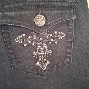 K&ouml;nnte beinhalten: Nahaufnahme einer schwarzen Jeans-Tasche mit einem dekorativen silbernen Kreuzdesign. Die Tasche hat einen silbernen Knopf mit dem Wort "Rock" und einem Stern. Das Kreuzdesign ist mit kleinen silbernen Nieten verziert.