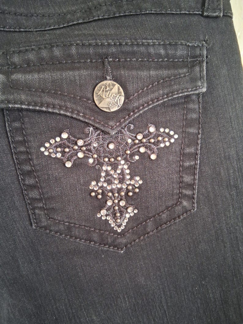 K&ouml;nnte beinhalten: Nahaufnahme einer schwarzen Jeans-Tasche mit einem dekorativen silbernen Kreuzdesign. Die Tasche hat einen silbernen Knopf mit dem Wort "Rock" und einem Stern. Das Kreuzdesign ist mit kleinen silbernen Nieten verziert.