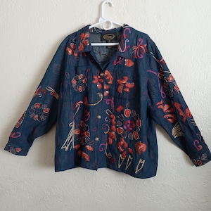 May include: A dark blue denim shirt with embroidered floral designs in shades of red, pink, and gold. The shirt has a button-up front and long sleeves.