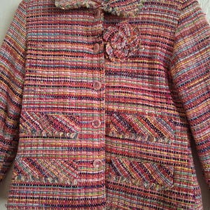 Op de afbeelding: Een veelkleurige tweedjas met een bloemversiering. De jas heeft een kraag, een knoopsluiting en twee voorzakken. De stof is geweven met roze, rode, gele en groene draden. De jas hangt aan een roze kledinghanger.