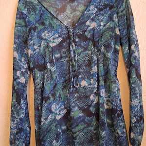 Könnte beinhalten: Eine langärmlige Bluse mit V-Ausschnitt und Schnürdetail. Die Bluse hat ein blaues, grünes und weißes Schlangenmuster. Das Kleidungsstück hängt an einem weißen Kleiderbügel.