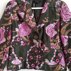 May include: A green embroidered blazer with pink and gold floral designs. The blazer has two buttons and a pocket on each side.
