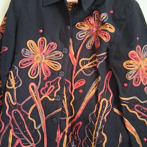 May include: Black jacket with colorful embroidered floral design. The jacket features a button-up front, a collar, and long sleeves. The floral embroidery includes red, orange, yellow, and white accents. The label reads "LANAGE".