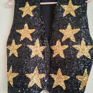 May include: Black sequined vest with gold star appliqués. The sleeveless vest has a V-neck and a single button fastening. The garment is suspended from a pink clothes hanger.