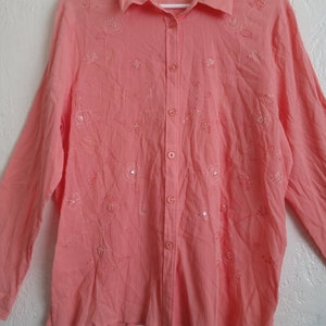 Può includere: Una camicia a maniche lunghe di colore rosa chiaro con bottoni, con motivi floreali ricamati e paillettes. La camicia ha un colletto e una chiusura con bottoni sul davanti.
