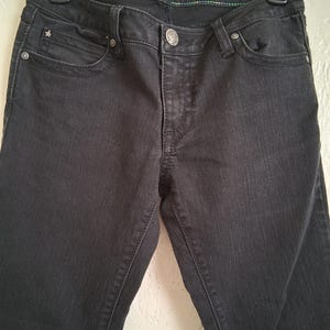 K&ouml;nnte beinhalten: Nahaufnahme einer schwarzen Jeans, die an einem schwarzen Kleiderb&uuml;gel h&auml;ngt. Die Jeans hat einen Knopfverschluss, Vorder- und Ges&auml;&szlig;taschen sowie eine kleine sternf&ouml;rmige Verzierung an der Vordertasche. Der Bund tr&auml;gt den Text "Rock 47 Wrangler".