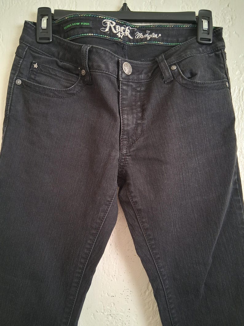 K&ouml;nnte beinhalten: Nahaufnahme einer schwarzen Jeans, die an einem schwarzen Kleiderb&uuml;gel h&auml;ngt. Die Jeans hat einen Knopfverschluss, Vorder- und Ges&auml;&szlig;taschen sowie eine kleine sternf&ouml;rmige Verzierung an der Vordertasche. Der Bund tr&auml;gt den Text "Rock 47 Wrangler".