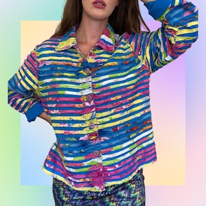 Puede incluir: Una camisa a rayas azules, rosas, amarillas y blancas con un estampado floral. La camisa tiene un frente abotonado y mangas largas.