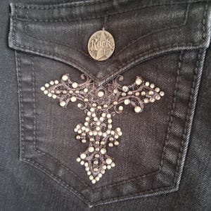 K&ouml;nnte beinhalten: Nahaufnahme einer schwarzen Jeans-Tasche mit einem dekorativen Kreuz-Design. Die Tasche hat einen silbernen Knopf mit dem Text "Rock 47". Das Kreuz-Design ist mit kleinen, funkelnden Strasssteinen verziert.