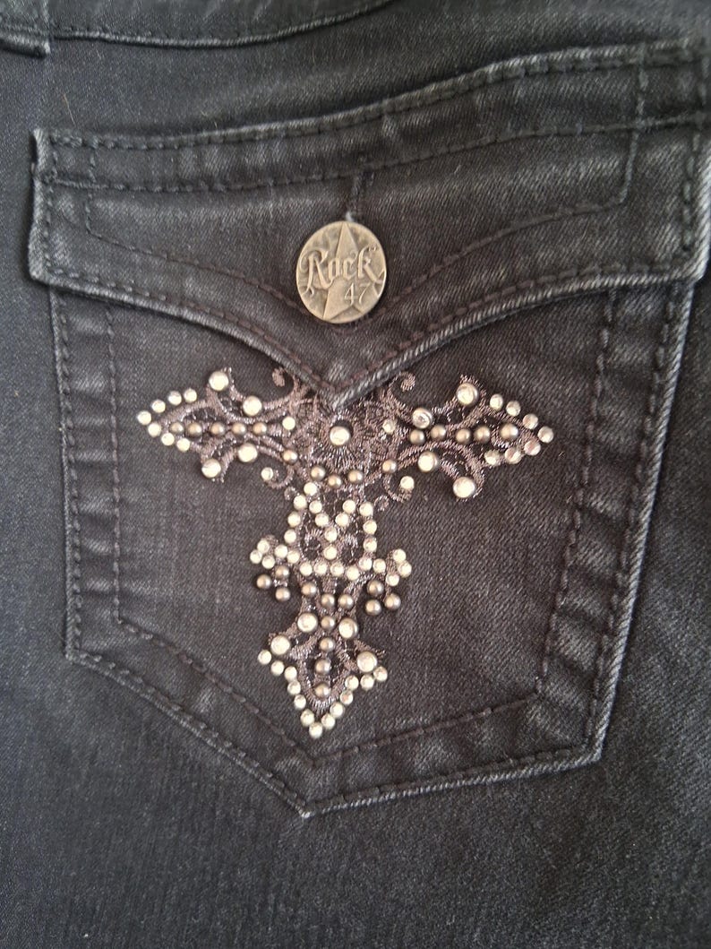 K&ouml;nnte beinhalten: Nahaufnahme einer schwarzen Jeans-Tasche mit einem dekorativen Kreuz-Design. Die Tasche hat einen silbernen Knopf mit dem Text "Rock 47". Das Kreuz-Design ist mit kleinen, funkelnden Strasssteinen verziert.