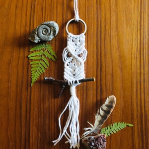 May include: White macrame owl keyring with brown beads for the eyes. The owl is hanging from a white cord with a metal ring. The owl is surrounded by natural elements like a fern, a small branch, a stone, and a feather.