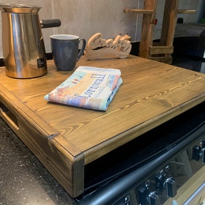 May include: A wooden tray with a brown finish sits on top of a black cooker. The tray has a handle on the side and is large enough to hold a coffee pot, a mug, and a book.