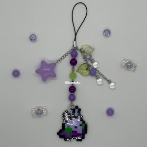 Puede incluir: Un dije pixelado morado y verde con una cadena, una estrella, cuentas y una cadena. El dije tiene un diseño pixelado de una criatura. El dije está rodeado de cuentas moradas y dijes en forma de caramelo. El texto "@iisankaku" es visible.